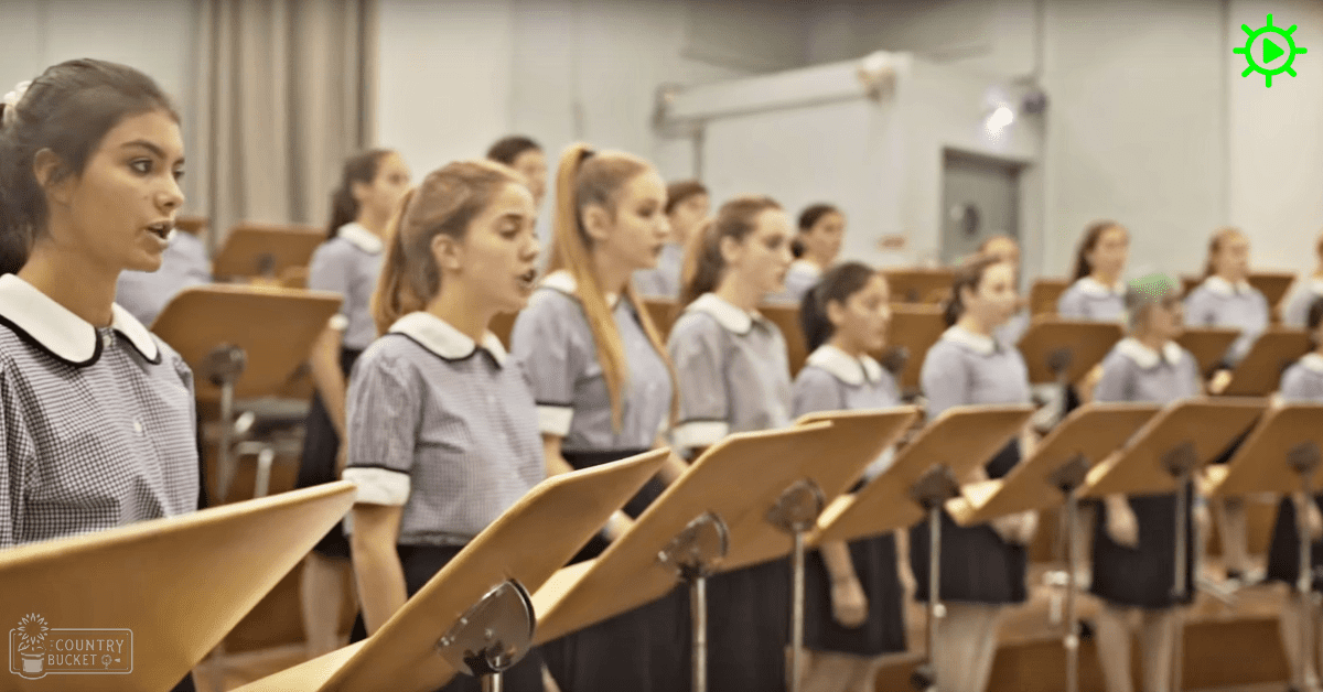 A group of young female adults in a choir setting at what appears to be a school auditorium, wearing uniforms with checkered shirts and dark skirts.
