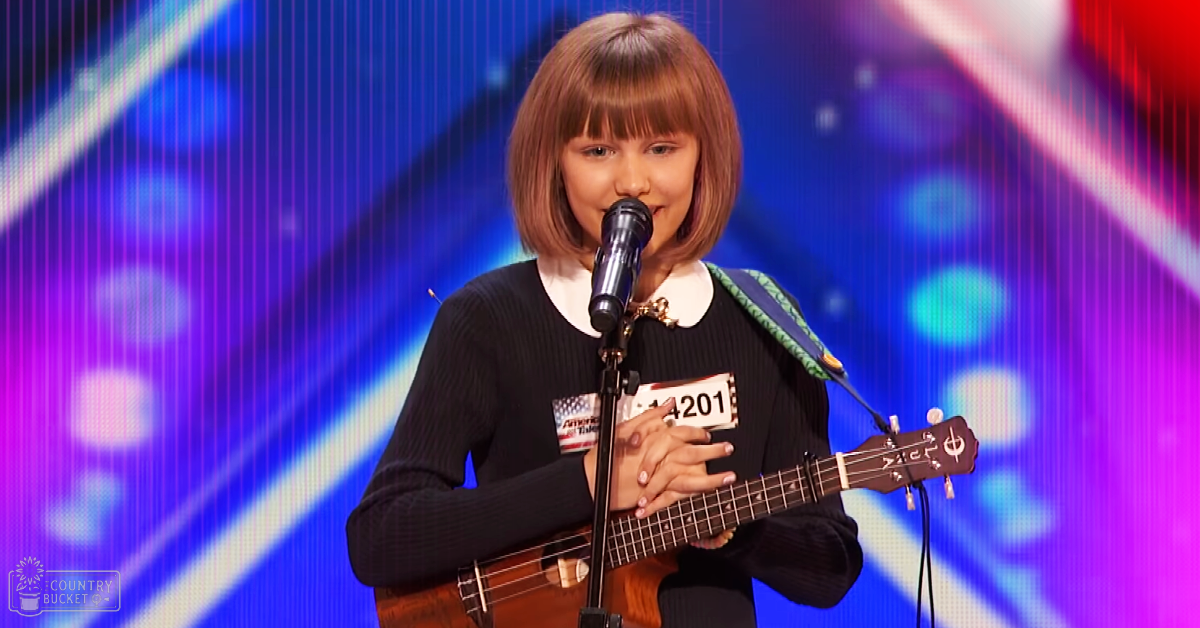 A young female child in a relaxed sweater with a Peter Pan collar, performing on an indoor stage with a microphone stand and ukulele, against a vibrant abstract background screen.