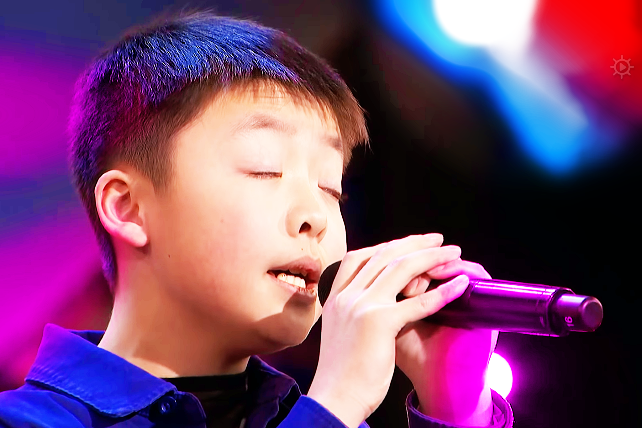 A young boy singing into a microphone at what appears to be a talent show or performance venue, wearing a blue button-up shirt.