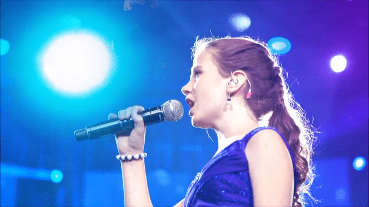 A young adult female performer in a fitted sleeveless dark blue dress with a scoop neckline, performing on stage under blue and purple lighting at an indoor concert venue.