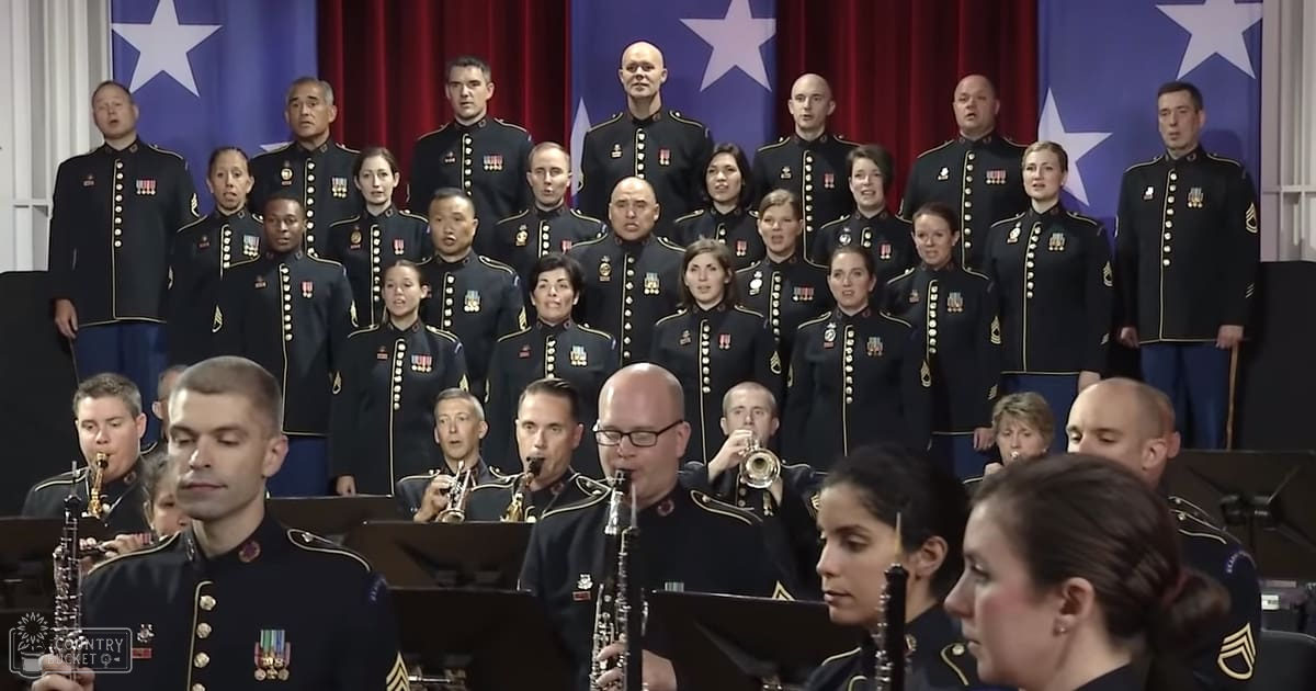 An adult male military choir performing with a formal stance in front of an American flag backdrop, surrounded by musical instruments under bright stage lighting.