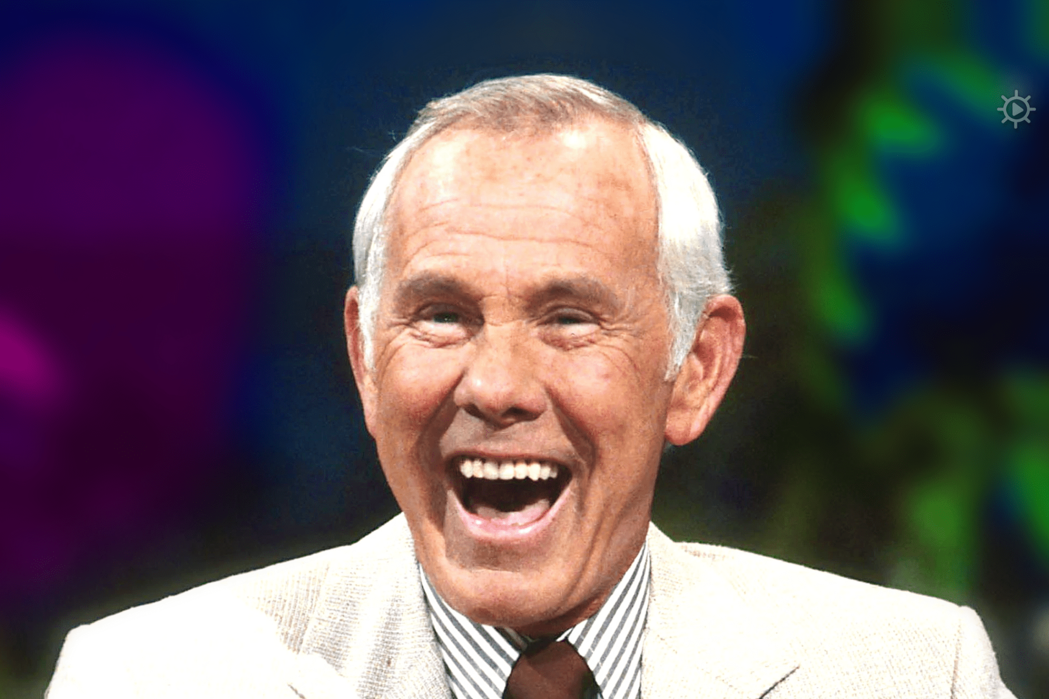 An older adult male in a studio setting, wearing a light-colored blazer over a striped shirt and dark tie.