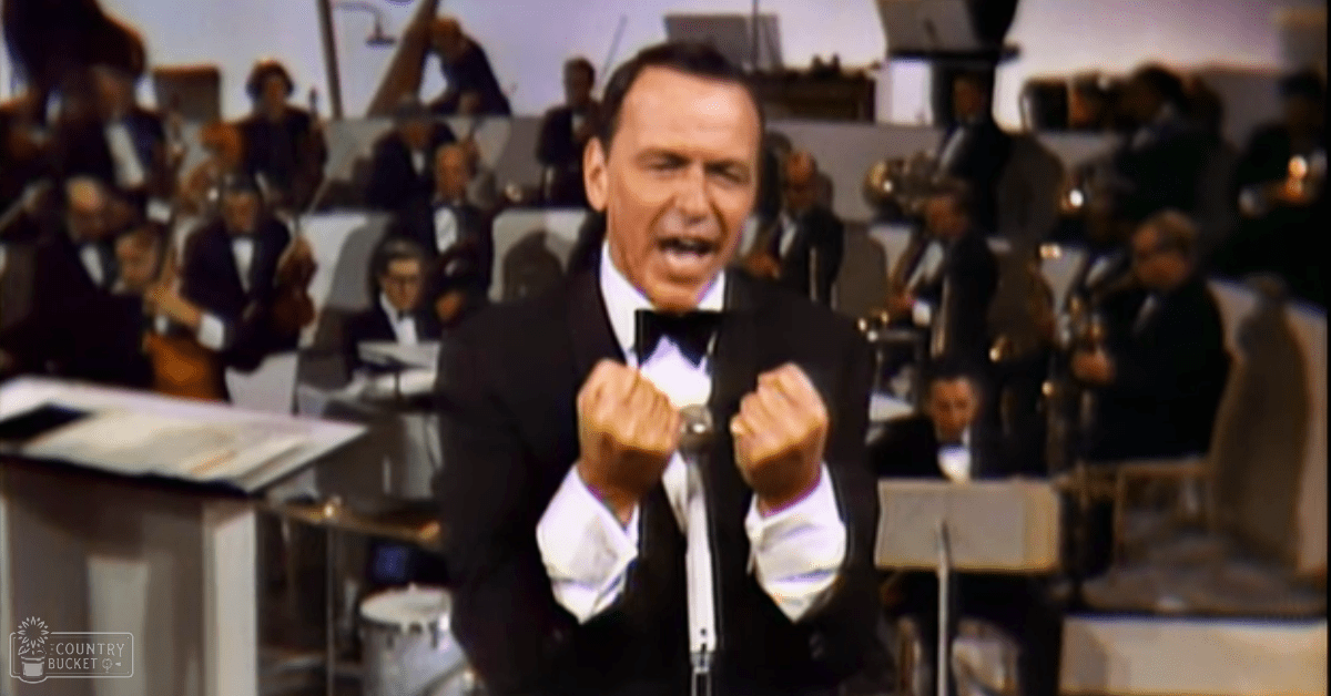 A middle-aged male performer in a black tuxedo stands before an orchestra during what appears to be a formal live performance or recording session, with rows of seated musicians and musical instruments visible in the background.