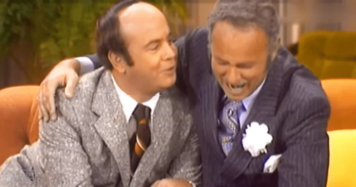 A comedic scene from an indoor television studio with two middle-aged men in formal attire, one wearing a textured blazer over a white shirt and tie, the other in a pinstriped suit jacket with a floral boutonniere. The setting includes a yellow couch and blurred greenery suggesting an outdoor backdrop.