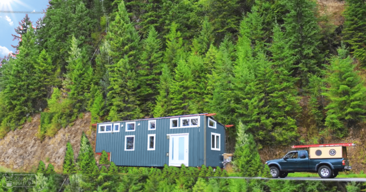 A blue pickup truck with camping gear parked near a tiny house amidst dense forested area with green foliage, possibly in spring or summer.
