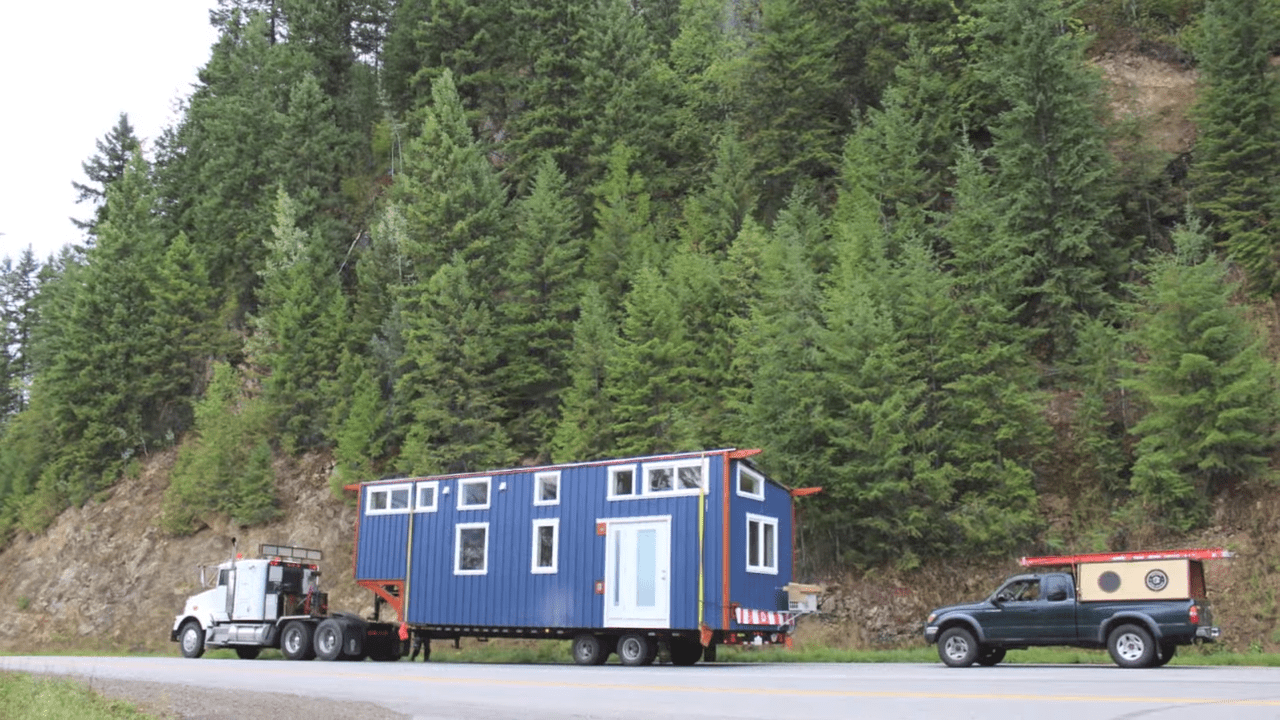 A semi-truck transporting a blue tiny house on a flatbed trailer in a forested area, with a smaller pickup truck nearby. The scene is outdoors, likely in spring or summer, with green foliage and clear skies.