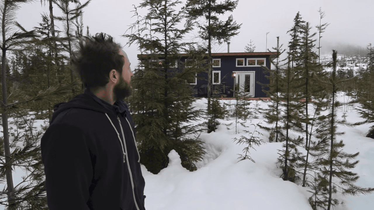 A young adult male wearing a dark hoodie in a snowy forested area with a small blue house surrounded by snow-covered trees.