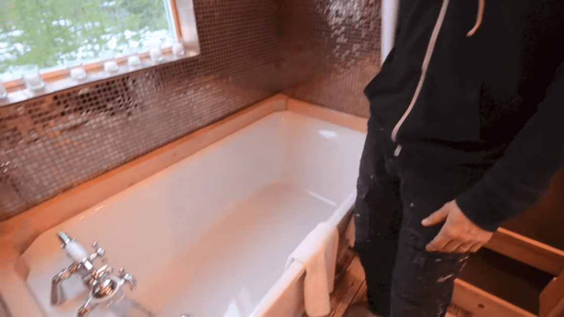 Bathroom renovation scene with adult male in black hoodie, mosaic tile walls, natural light, unfinished walls.