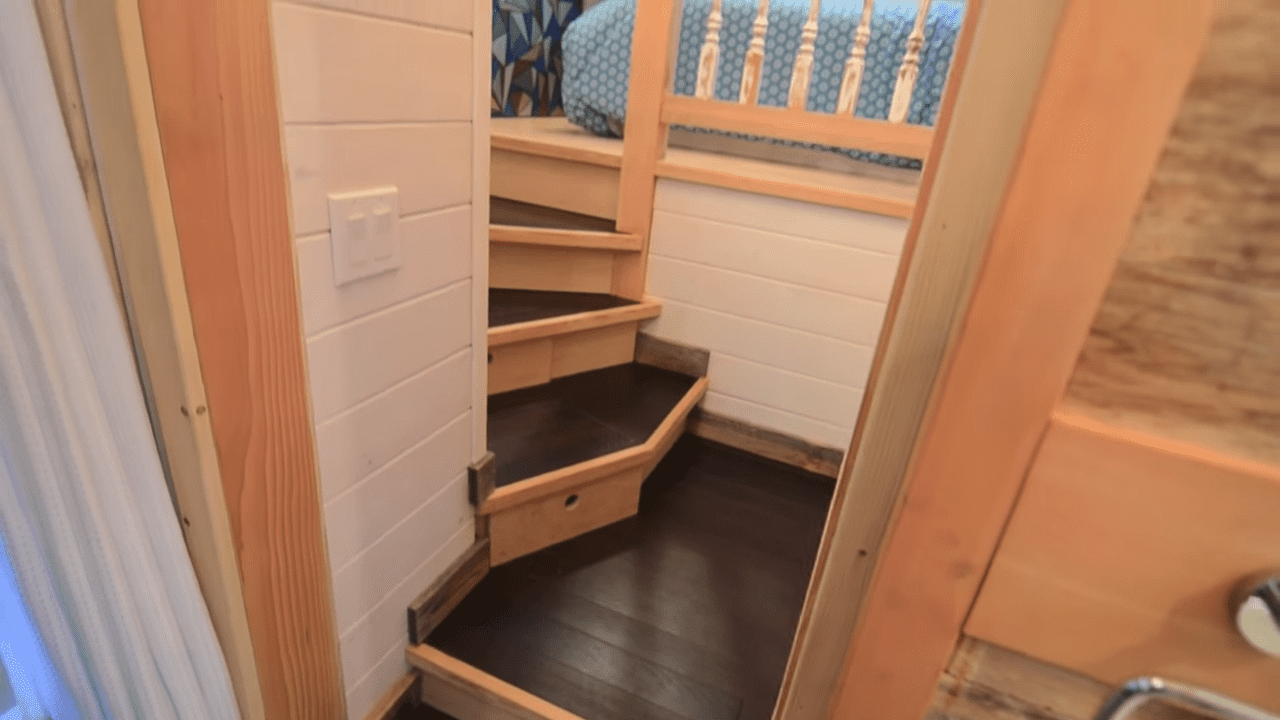 Wooden staircase leading to loft bed in compact living space with white shiplap walls, small electrical outlet visible, natural light from window, daytime scene.