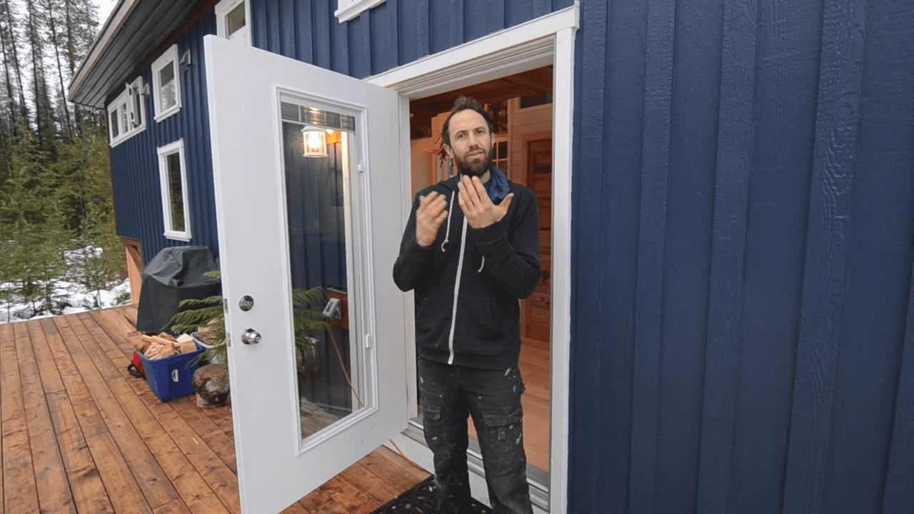 Adult male wearing a black zip-up hoodie in front of a blue cabin with snow-covered trees and wooden deck.
