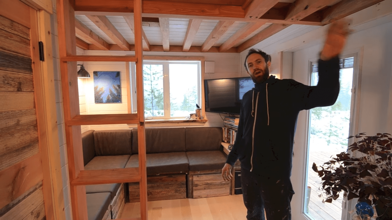 An adult male in a cozy living space with wooden beams, wearing a dark blue zip-up hoodie indoors during winter with natural light and snow visible through the window.