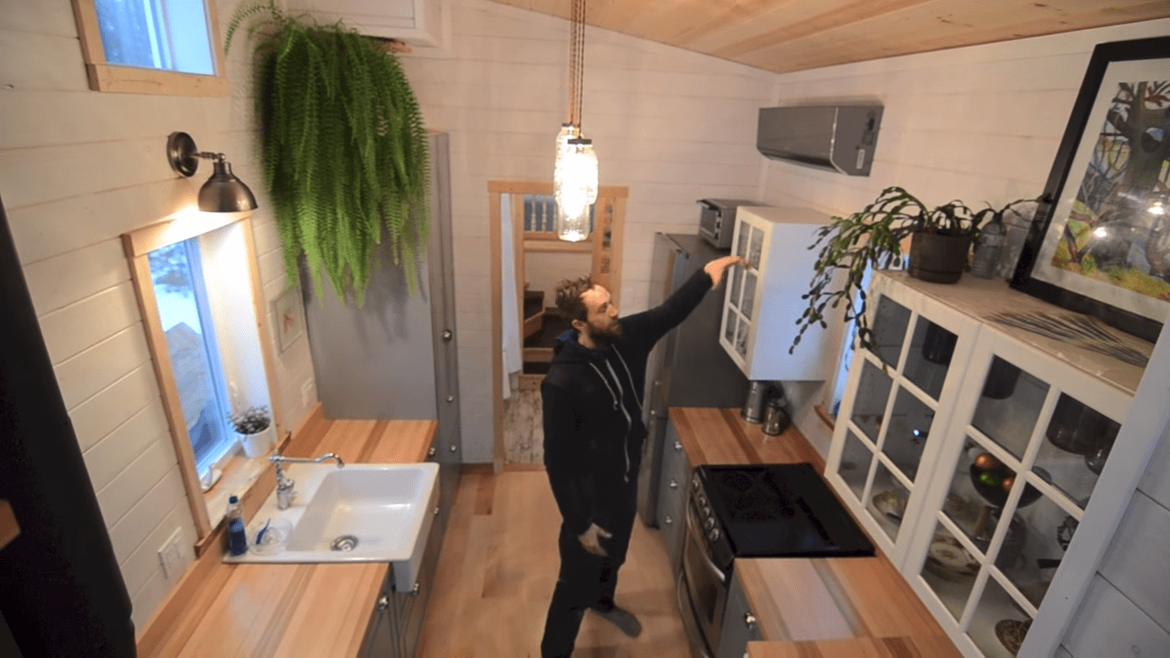 An adult male in a kitchen within a tiny house, surrounded by white cabinets with glass doors, wooden countertops, a sink, stove, and hanging light fixture. Natural light illuminates the scene.