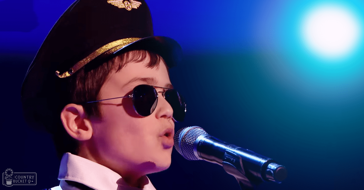A child in a performance setting, wearing a white collared shirt with black cap with gold trim, stage lighting with blue and white hues.