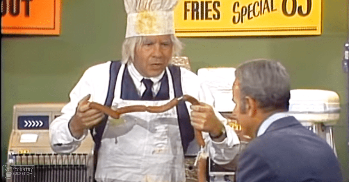 Middle-aged man in a retro-style fast-food setting, wearing a white chef's hat with a gold emblem and an apron over a vest or suit jacket. Another man is dressed in a dark blue modern suit.