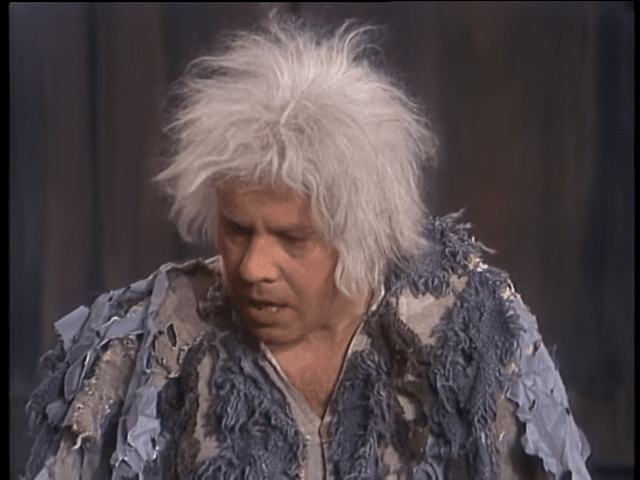 An older adult woman in a theatrical setting, wearing a loose, flowing blue-gray ruffled jacket with darker patterns, standing against wooden paneling.