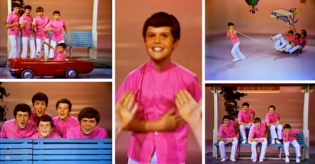 A group of young male children in bright pink shirts perform or pose on a stage designed to resemble an outdoor bus stop area with a blue bench labeled 'BUS STOP'.