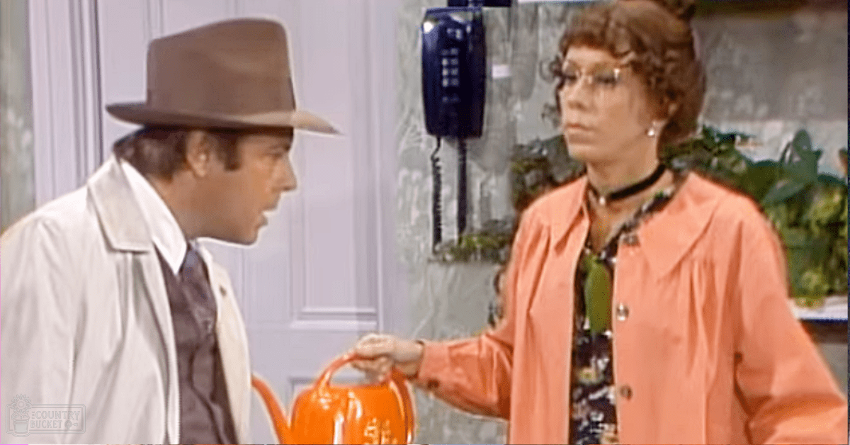 An adult male wearing a light-colored trench coat with high collar, dark shirt, and brown fedora-style hat indoors; an adult female in a peach-colored jacket over patterned blouse with green accents and black choker necklace, surrounded by vintage decor including a telephone and plants.