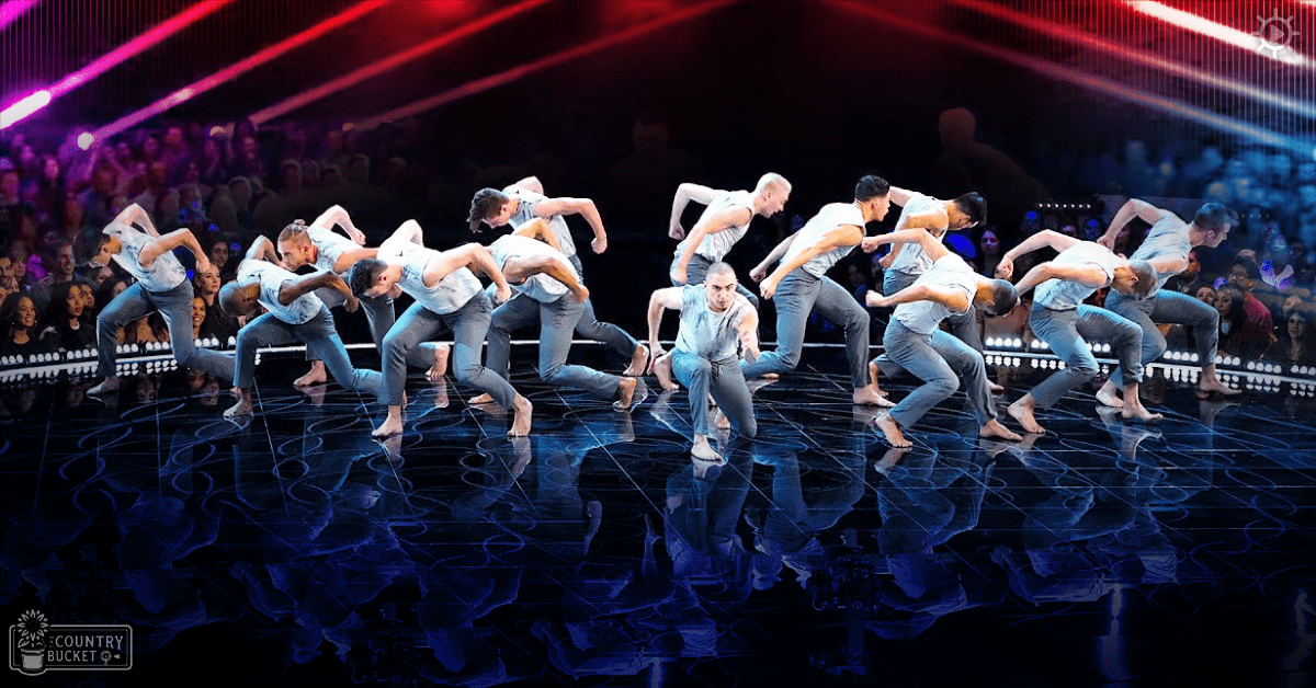 A young adult male dance troupe performing energetically on stage at an indoor event with bright red and purple lights and reflective flooring.