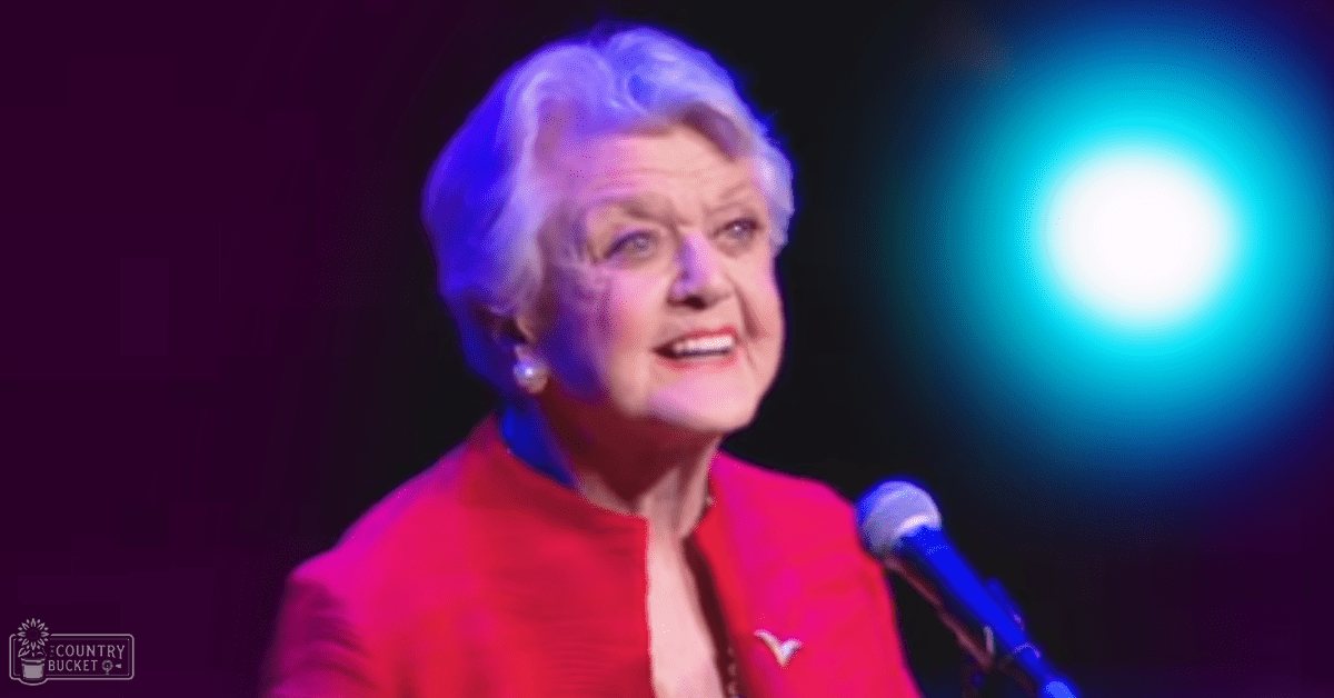 A middle-aged woman in a fitted red blazer with pearl earrings stands on stage, illuminated by bright blue light against a dark background.