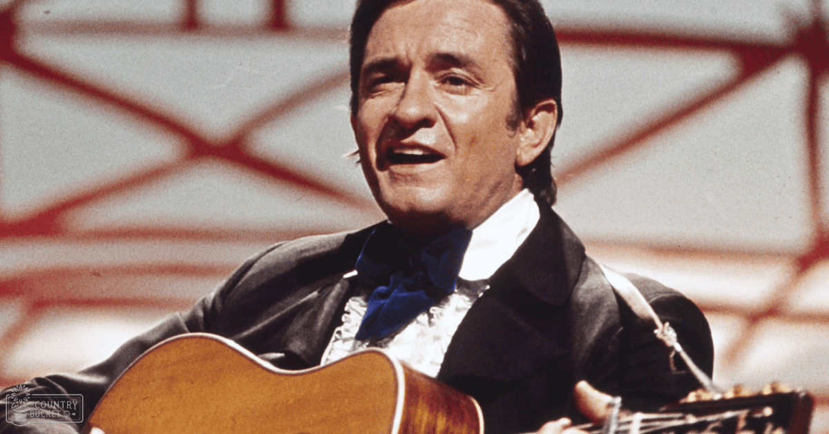 A middle-aged male performer in a dark suit with a white shirt and blue bow tie, standing on an indoor stage or performance area with a blurred red-lined background.
