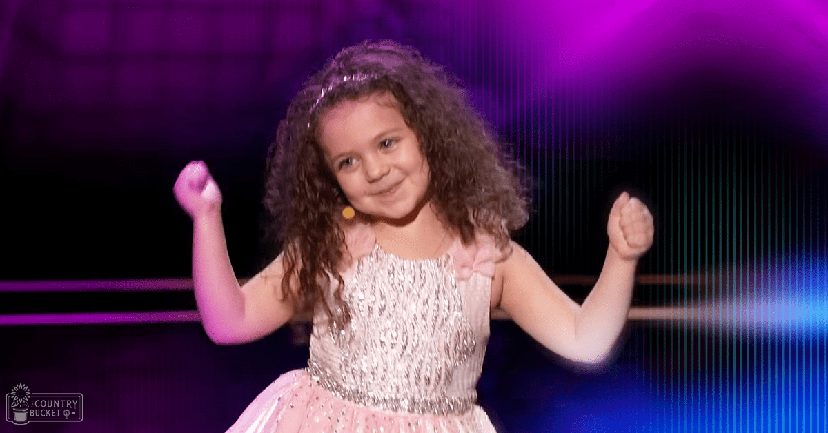 A young child with curly hair wearing a sparkly pink dress holds a microphone, possibly performing on stage.