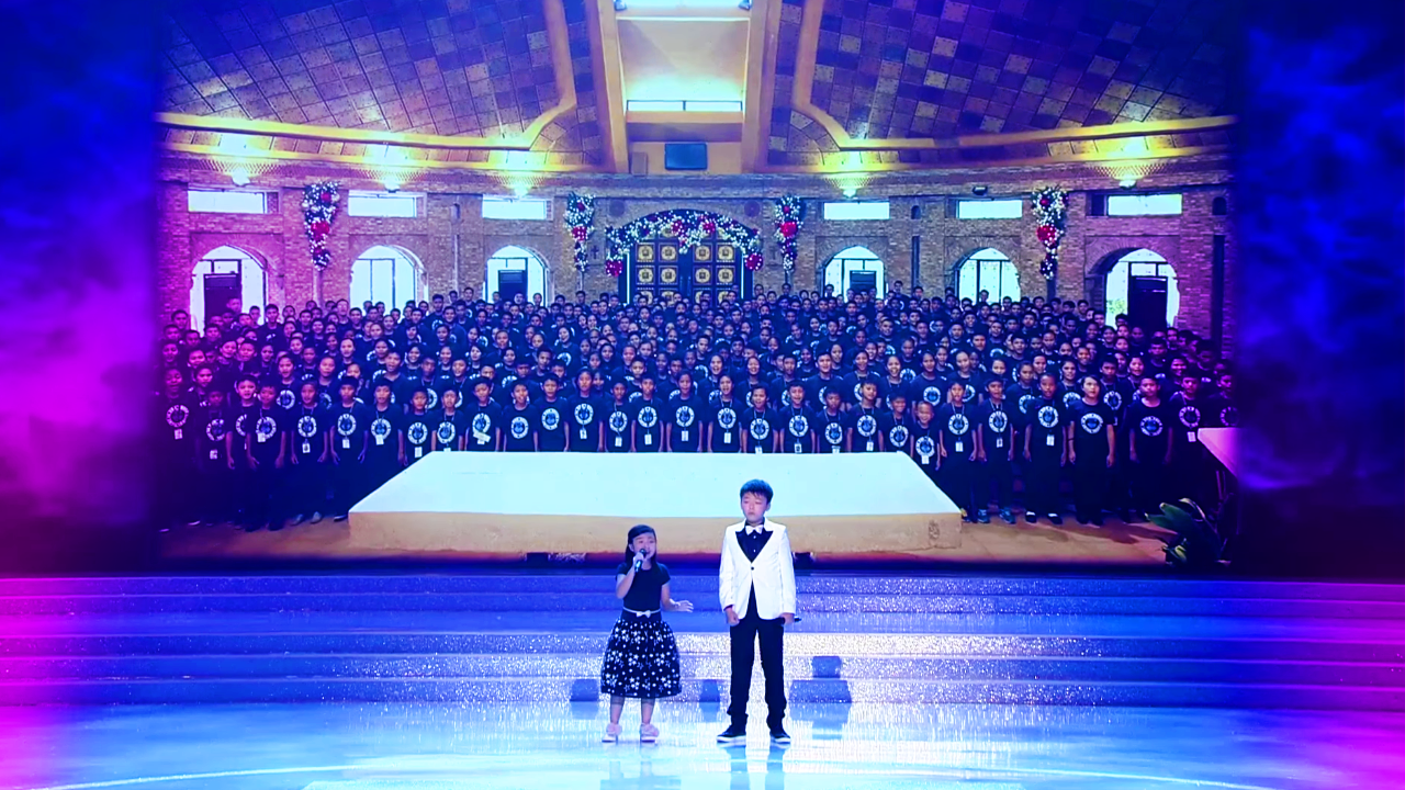Children perform at an indoor venue with brick walls and stage lighting, with a toddler in a black polka-dot dress holding a microphone.
