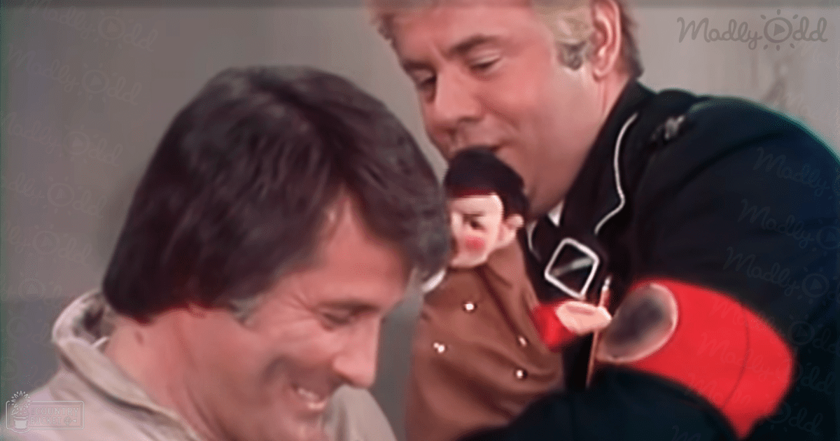 Middle-aged man in a light-colored hoodie or jacket with high collar indoors; young adult male in uniform-like garment with white trim and buttons behind him, older adult man in dark jacket with red accents on sleeve further back.