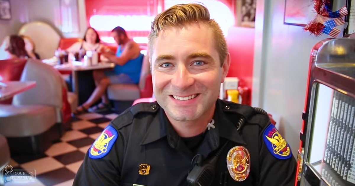 A police officer in a dark blue uniform stands at the counter of a diner, while two women sit at a booth wearing sleeveless dresses. The background includes an American flag decoration and a cash register.
