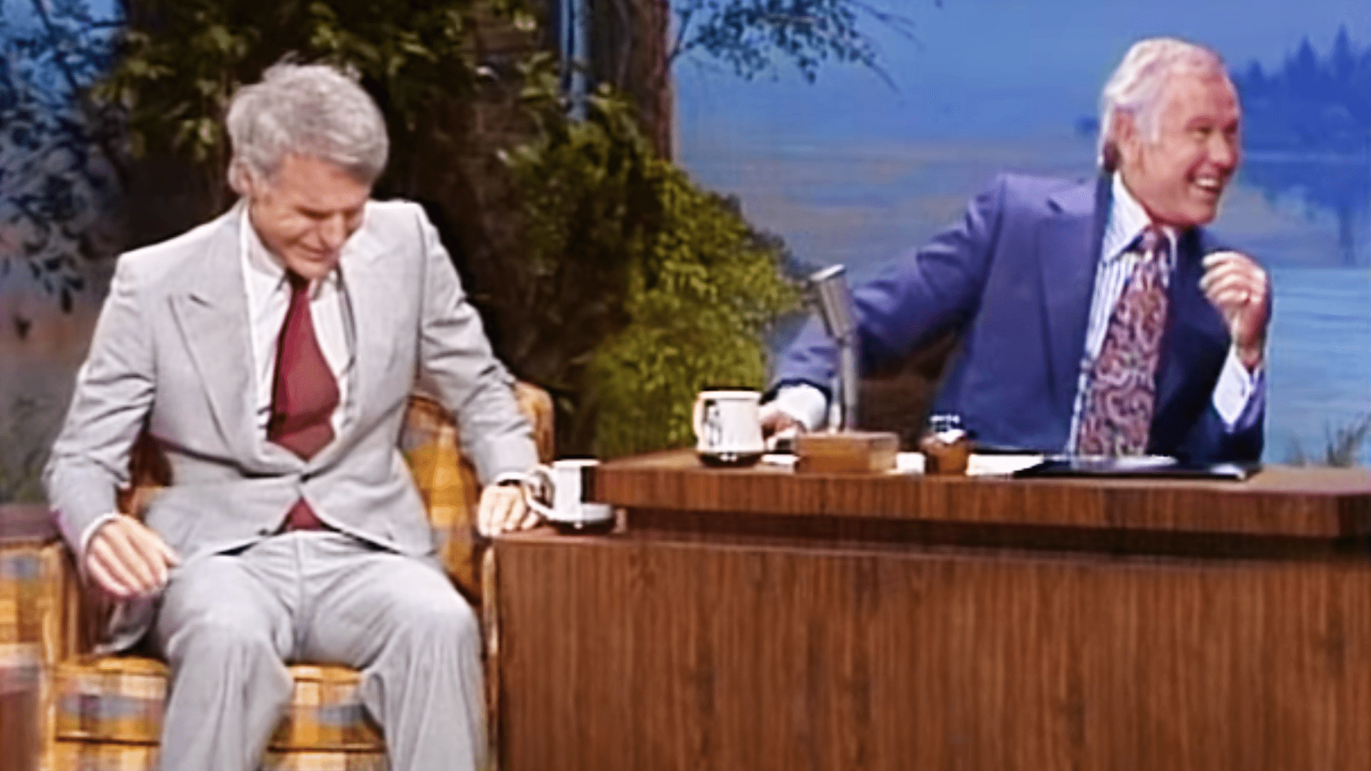 Two older adult men seated at a wooden desk with microphones and coffee mugs, against a scenic backdrop of trees and water in what appears to be a television studio designed for talk shows.