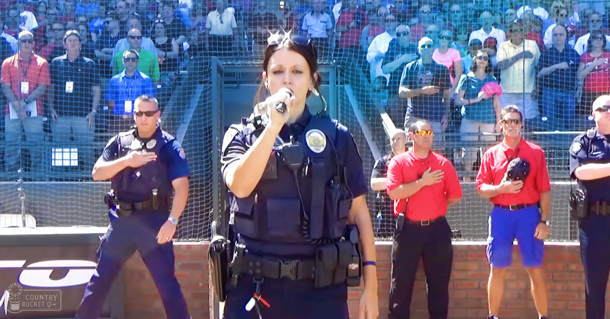 Adult female police officer singing into a microphone at an outdoor baseball stadium during spring or summer, with diverse spectators in the background.