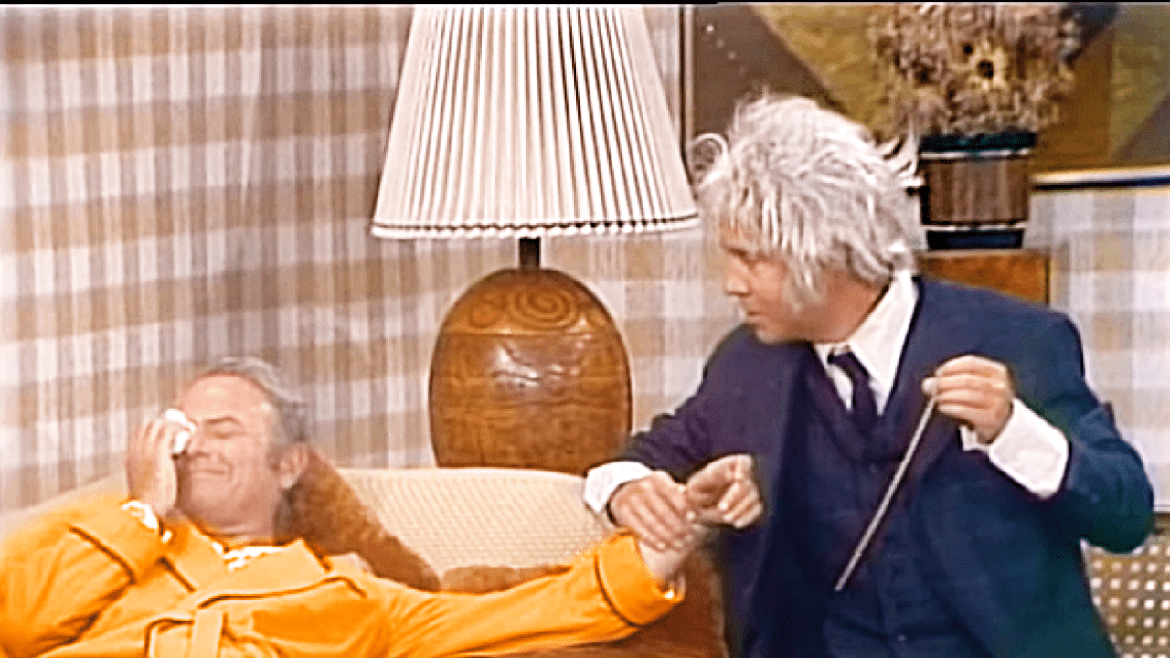 A comedic scene from a television show or film set in an indoor living room with mid-century modern decor, with a middle-aged male in a bright yellow robe and an older adult male in a dark blue suit.