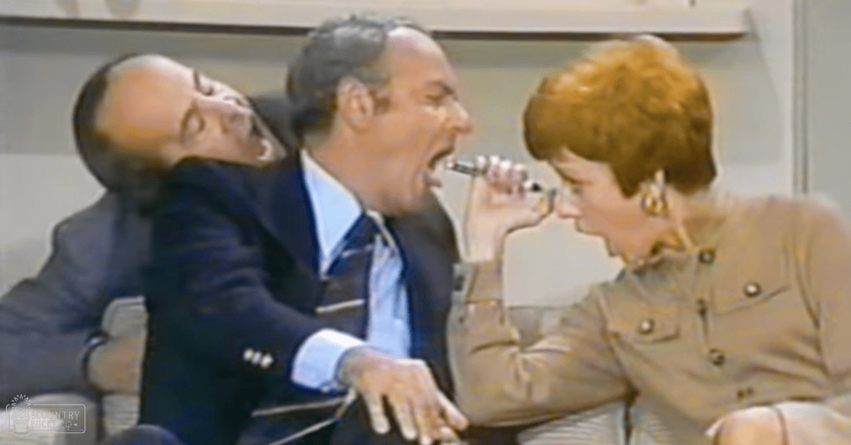 Three adults in a comedic indoor studio setting, reacting to something humorous; one holds a microphone while another laughs.