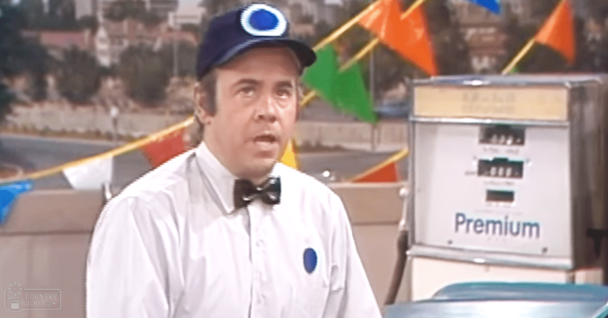 A middle-aged male wearing a white shirt with a bow tie and dark blue cap stands at what looks like a gas station surrounded by outdoor elements such as trees, colorful flags, and part of a car. The setting suggests an outdoor location during daytime.
