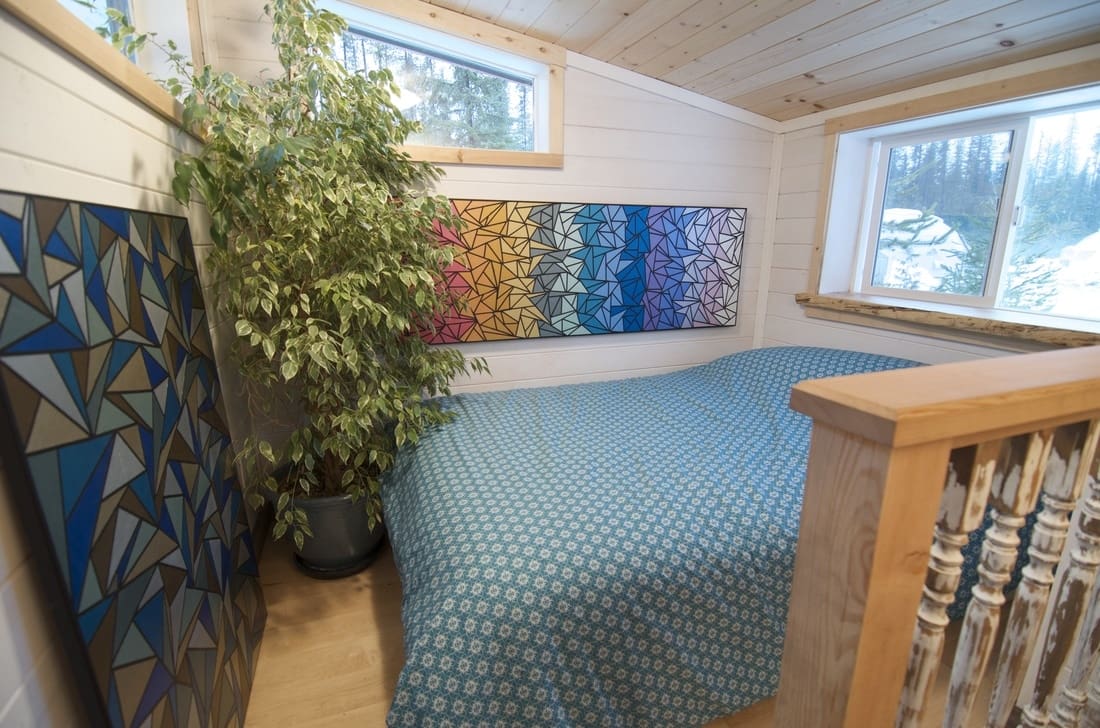 A cozy bedroom with blue patterned bedding, a large potted plant, colorful geometric artwork, white paneling walls, two windows showing snowy trees outside during daytime.