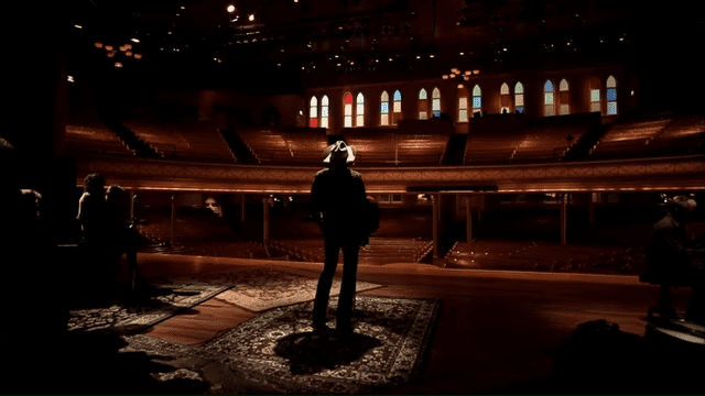 An adult male stands indoors, possibly during a performance or rehearsal, in an auditorium with rows of empty seats and arched windows lit from behind.