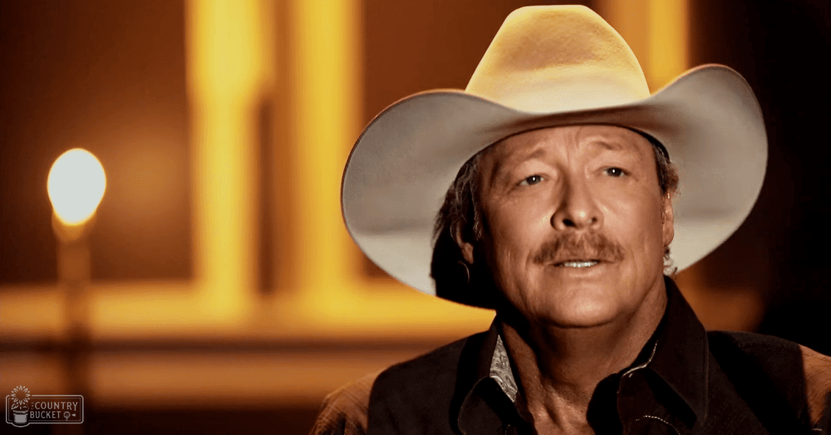 An older adult male wearing a wide-brimmed cowboy hat and dark-colored shirt stands against an indoor setting with warm lighting and architectural elements.