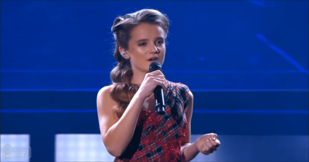 A young adult female singer in a red dress with black plaid patterns performs on stage under blue lighting, holding a microphone.