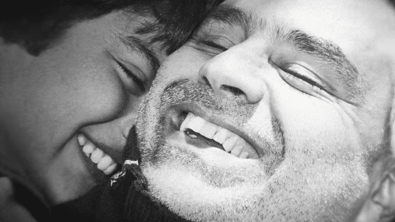 Close-up interaction between two individuals, one young adult with dark hair smiling, older male with beard laughing, indoors, no clear season or time of day, black and white image.