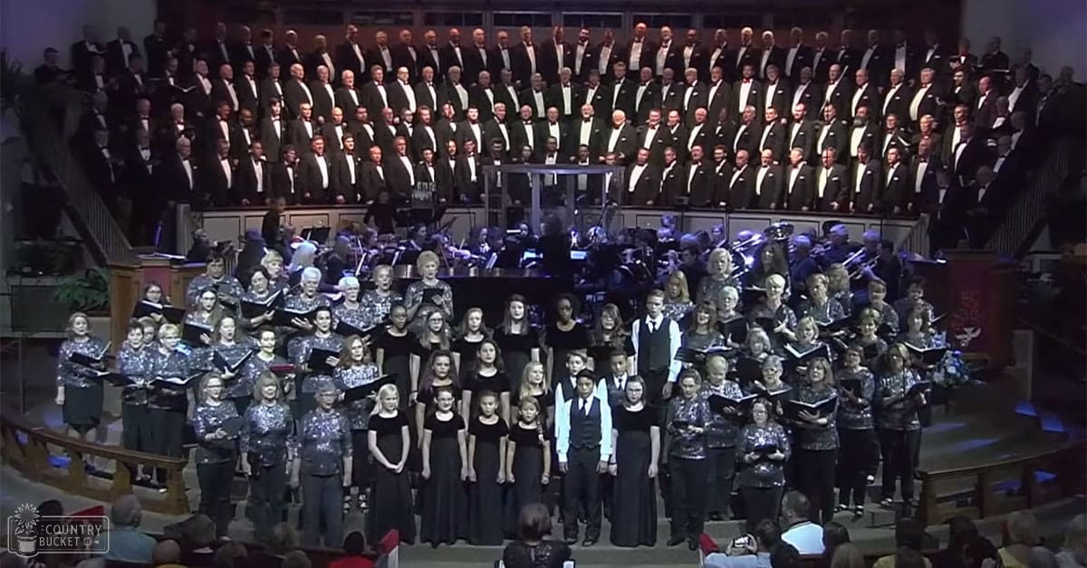 A choir performance in a large auditorium with adults in formal black tuxedos and women in shimmering dark dresses, likely during a holiday season.