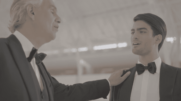 An older adult male in a dark suit jacket adjusts a young adult's bow tie indoors under bright lighting, surrounded by blurred furniture.