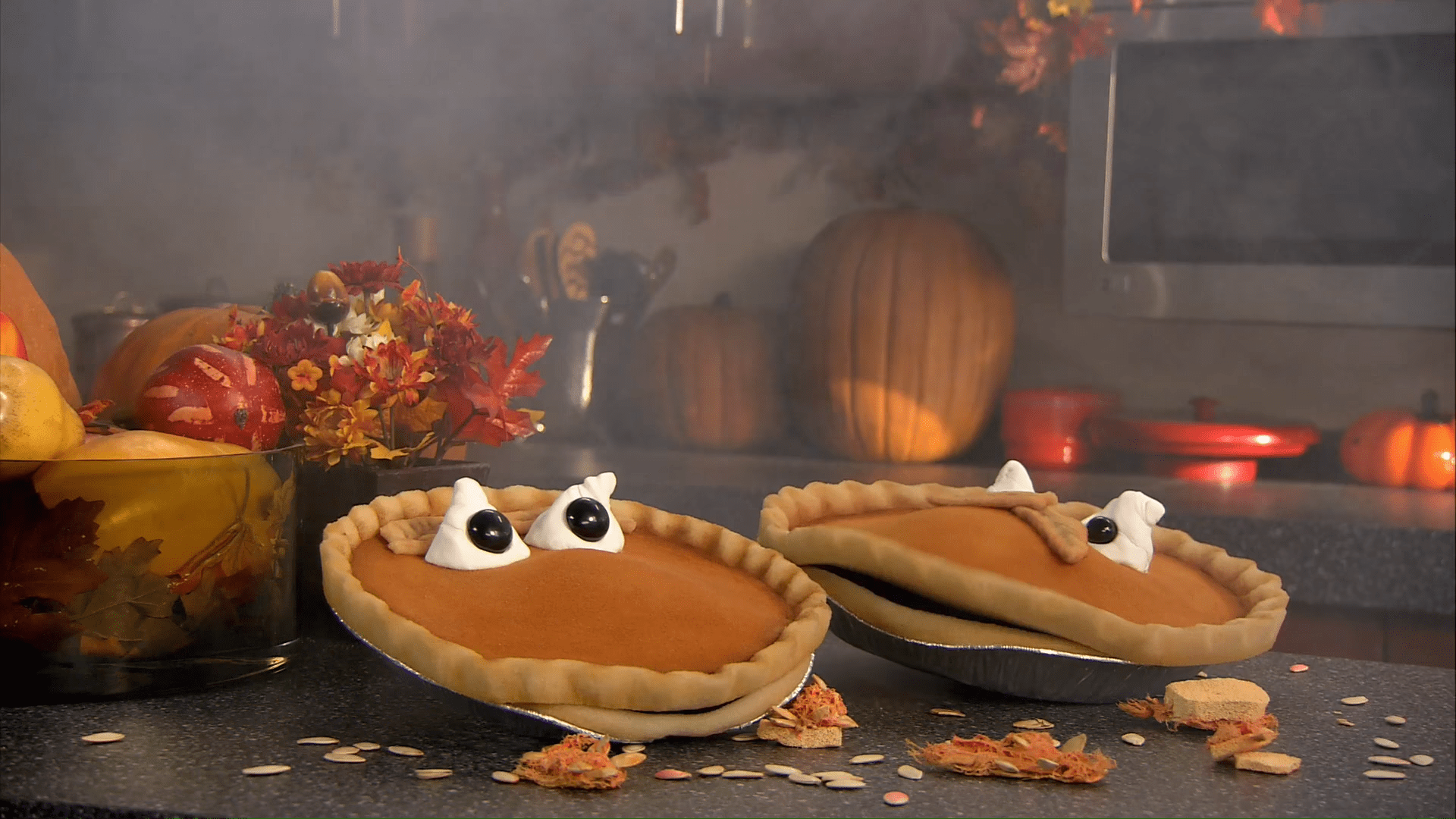 Halloween-themed dessert setup in a kitchen or dining area with autumnal decorations, including pumpkin pies, scattered pumpkin seeds, and fall foliage.