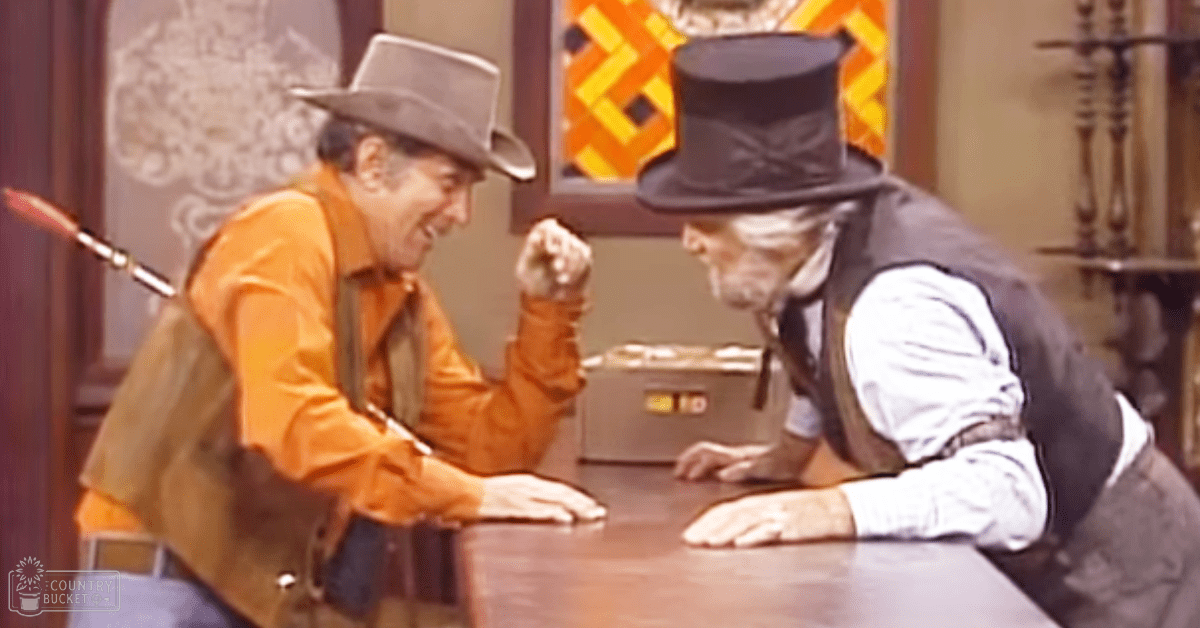 A scene from a Western movie or TV show, set in the 19th century with two men in period attire, one holding a knife and leaning on a table.