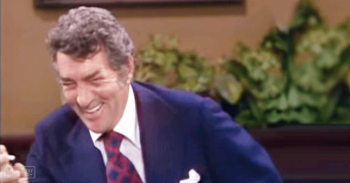 A middle-aged man wearing a mid-20th-century dark blue suit jacket, white dress shirt, and red tie with black spots sits indoors against a backdrop of artificial greenery under bright studio lighting.