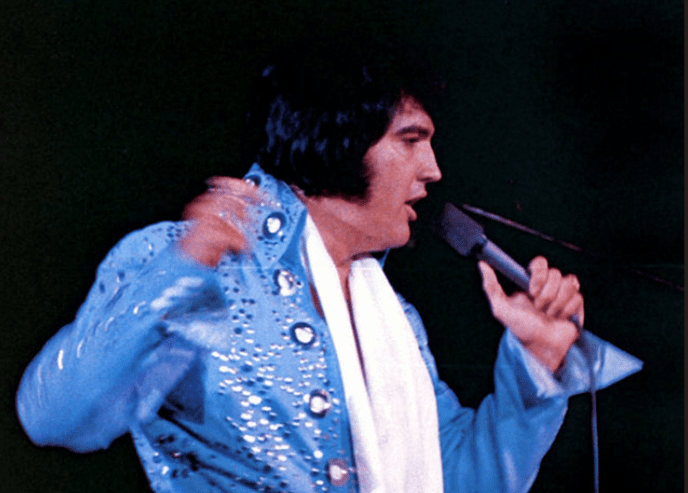 A male performer in a blue sequined 1970s or early 80s stage costume, holding a microphone on an indoor stage with dim lighting.