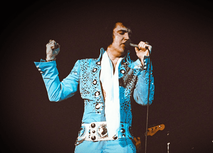 An adult male performer in a blue sequined 1970s-style jacket with white accents, holding a microphone on stage under dim lighting.