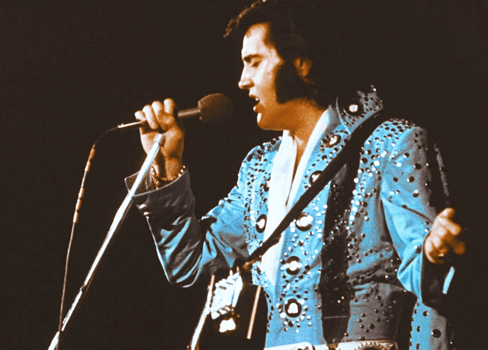 An adult male performer in a concert setting, wearing a blue sequined 1970s-style jacket with black lapels and embellishments, holding a guitar strap over his shoulder.