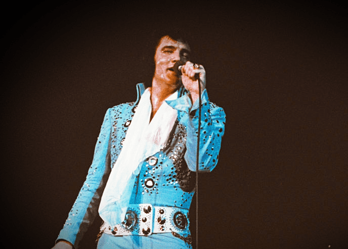An adult male performer in a light blue, intricately sequined jumpsuit with a high collar on stage.