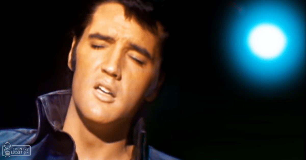 An adult male in an indoor setting, wearing a relaxed fit open-collared dark blue shirt with mid-20th-century style and dimly lit by spotlight.