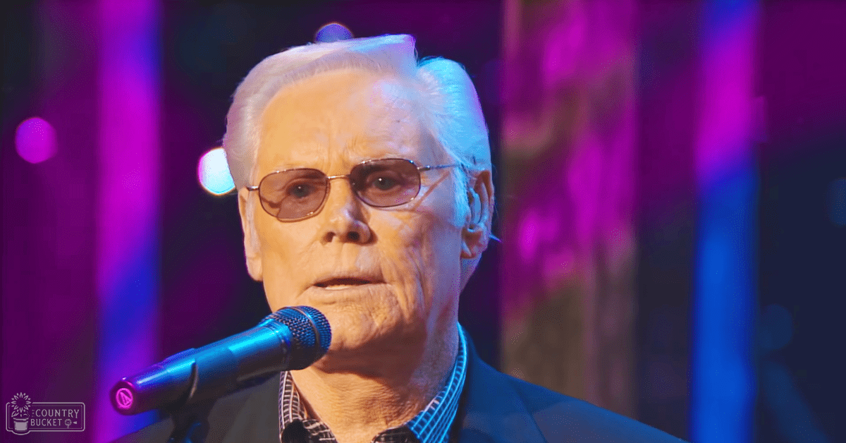 An older adult male in a dark suit jacket and collared shirt with subtle checkered pattern stands on a stage under vibrant purple and blue lighting, likely during a performance.