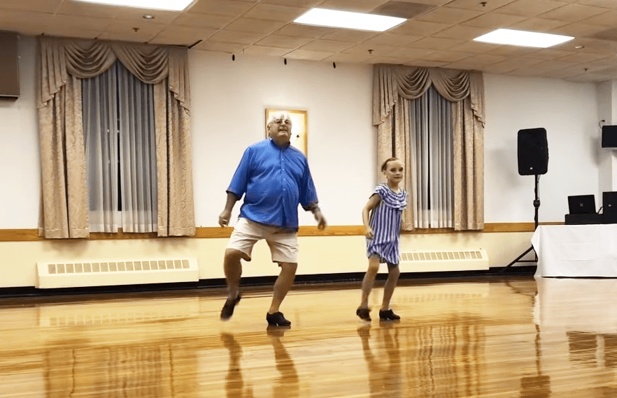 An adult male dancer and a young girl in an indoor dance studio, wearing a blue button-up shirt and striped dress respectively.